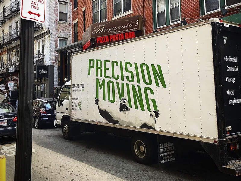 Precision Moving truck parked on a city street, with a pizza restaurant in the background.