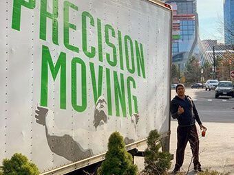 Man gives thumbs-up next to a Precision Moving truck. Green lettering, outdoor setting with buildings.
