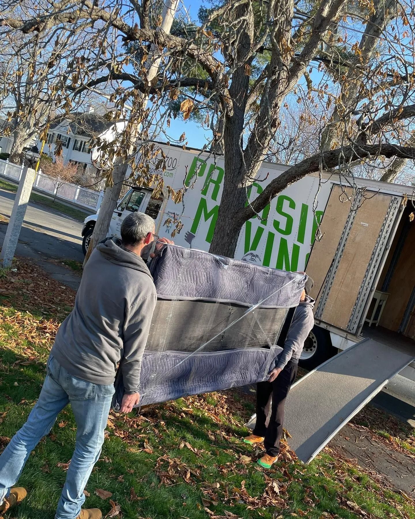 Two people carrying a wrapped mattress toward a moving truck on a lawn, beneath a tree.