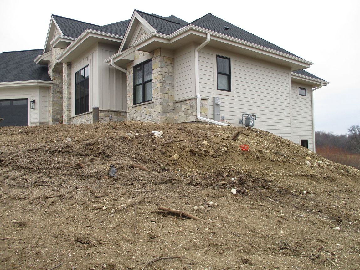 A two-story light-colored house with stone accents, dark shingles, and black window frames on an unfinished dirt slope.