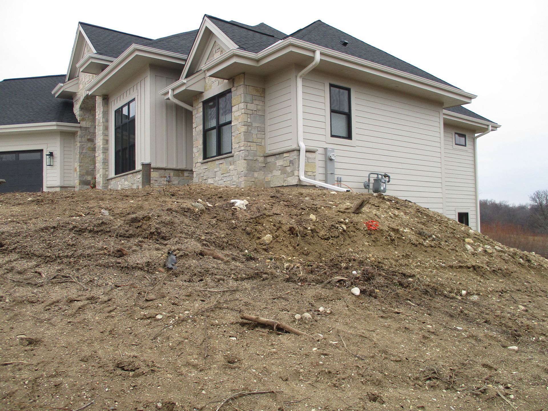 A two-story light-colored house with stone accents, dark shingles, and black window frames on an unfinished dirt slope.