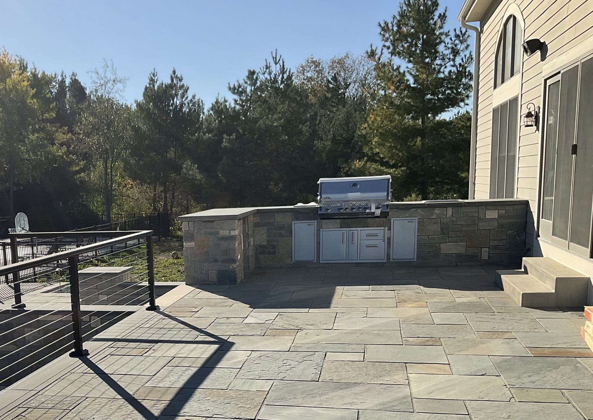 Outdoor stone patio featuring a built-in stainless steel grill set into a stone counter, beside a house with steps.