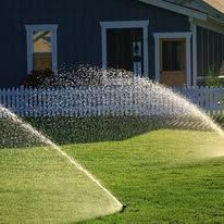 A lawn sprinkler is spraying water on a lush green lawn in front of a house.