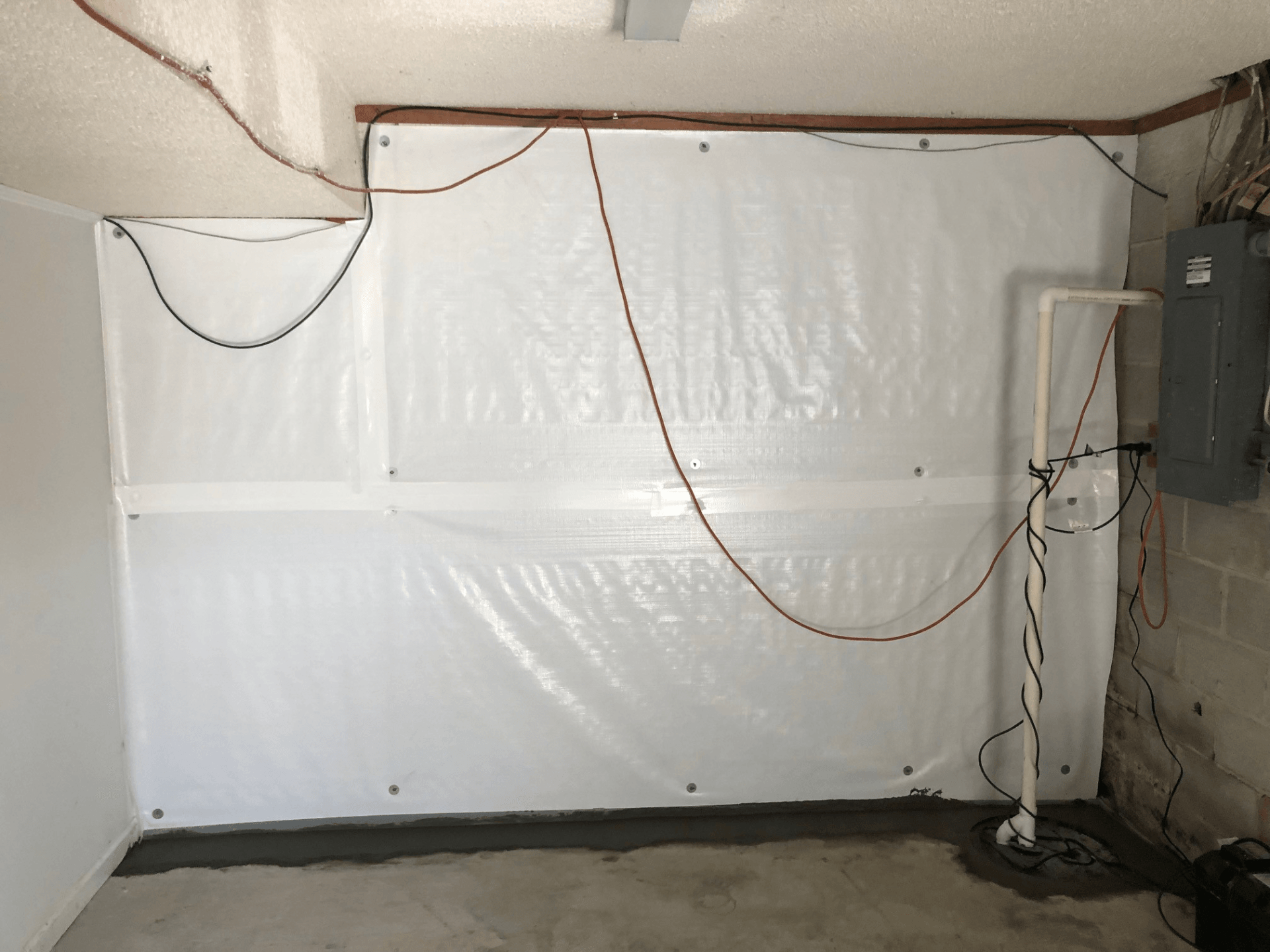 White plastic sheeting covers a wall in a basement, with electrical wires and a water pipe attached.