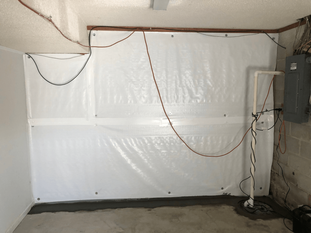 White plastic sheeting covers a basement wall. Wires and a pipe run along the wall. A cement floor is in the foreground.