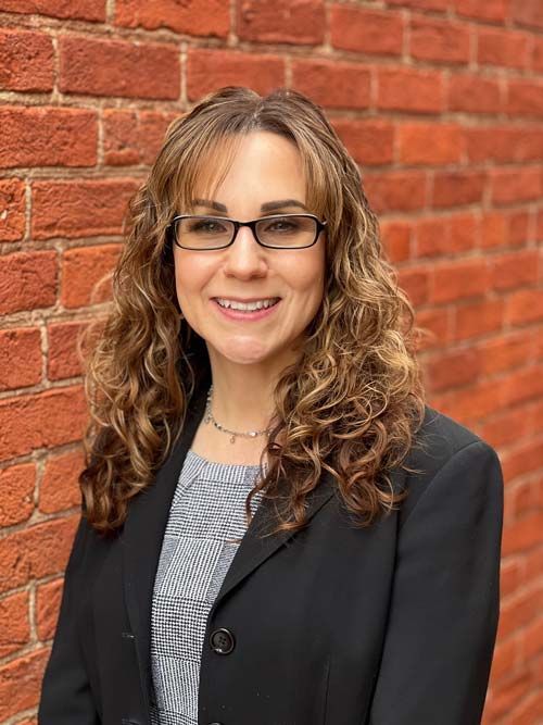 A woman wearing glasses and a black jacket is standing in front of a brick wall.