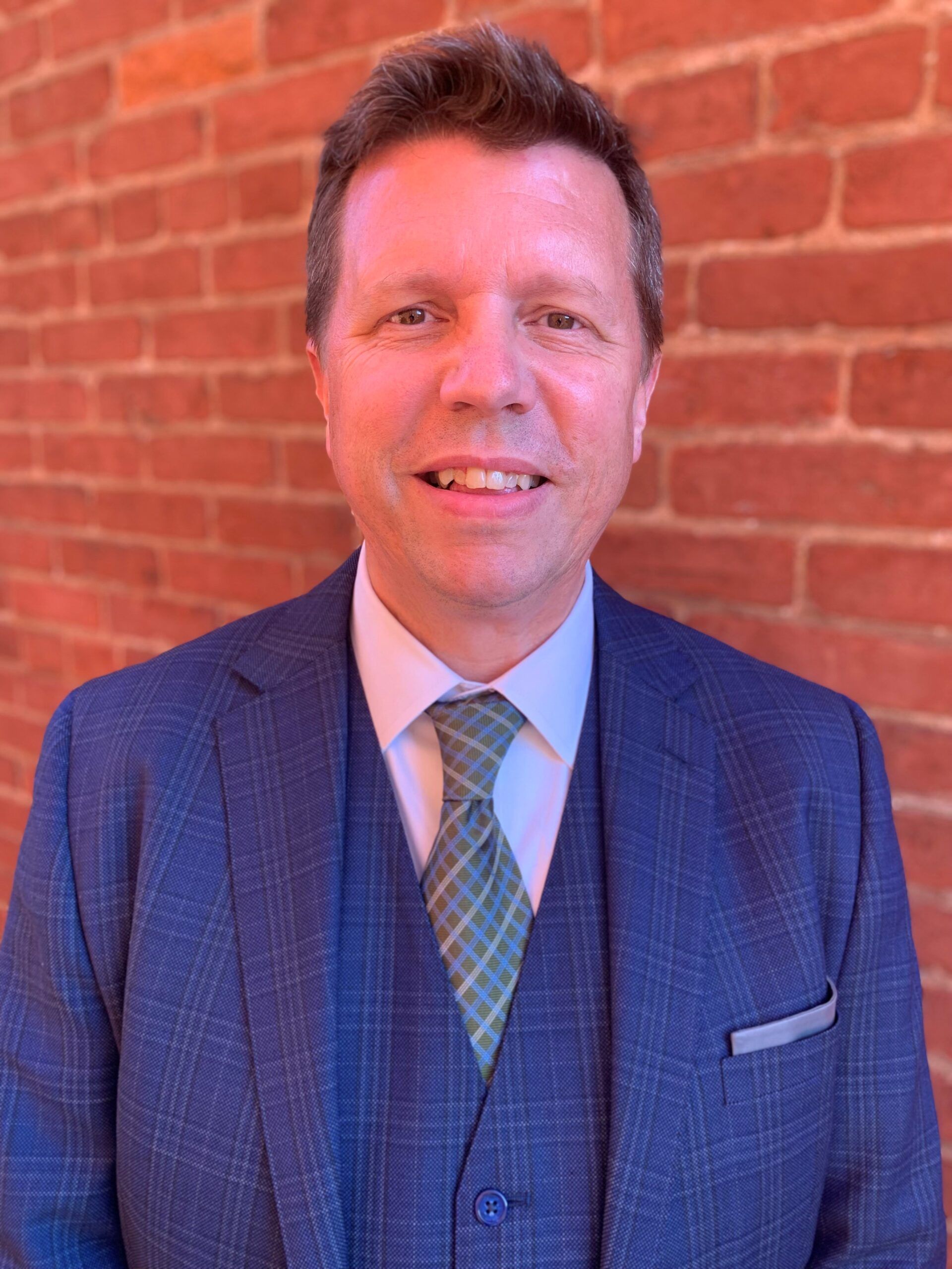 A man in a suit and tie is smiling in front of a brick wall.