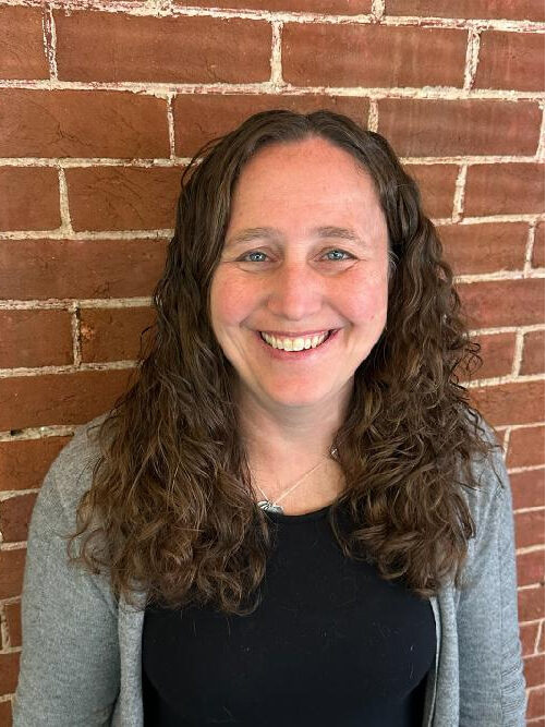 A woman with curly hair is smiling in front of a brick wall.