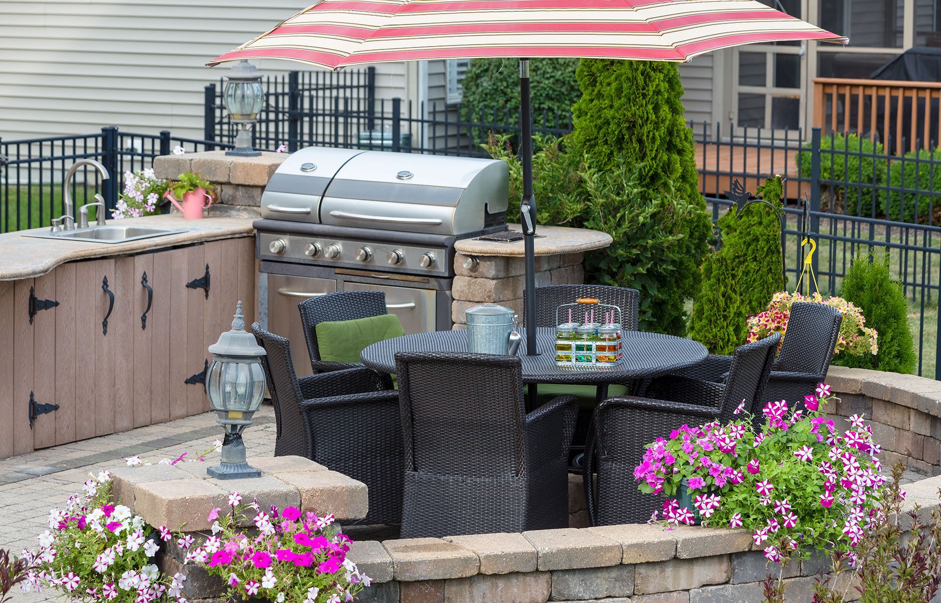 Outdoor kitchen with grill, dining table, and umbrella, set in a patio with brick accents and flower beds.