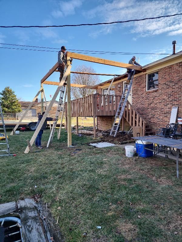 Construction workers building a wooden deck cover in a backyard, using ladders and power tools. Sunny day.
