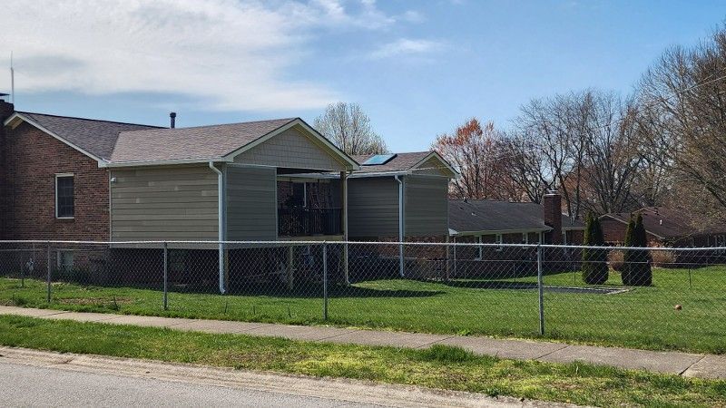 Green-sided houses with brown roofs behind a chain-link fence on a grassy lawn under a blue sky.