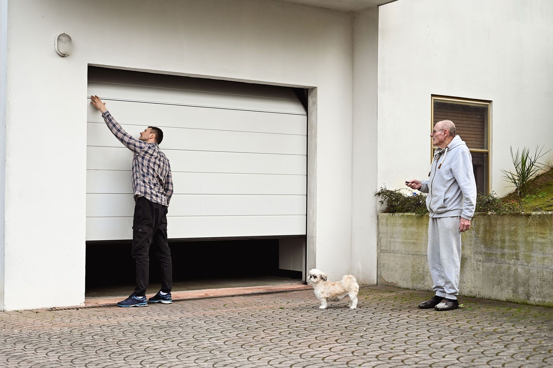 Two men and small dog near partially open garage door; one man reaches up.