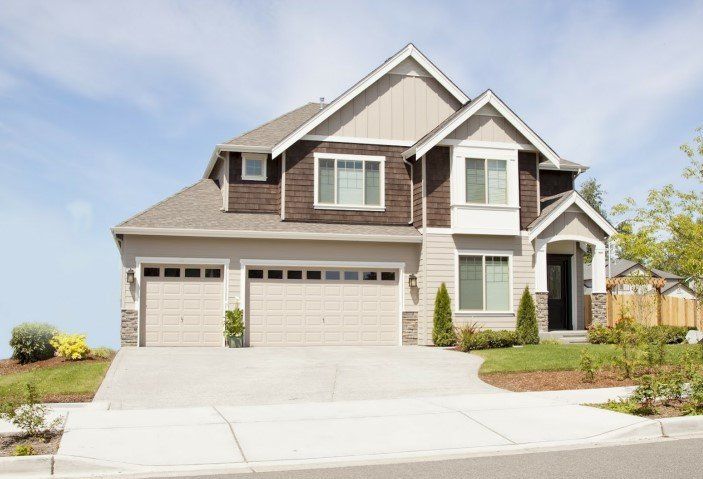 Two-story house with tan garage doors, light-colored siding, brown shingles, and a concrete driveway.