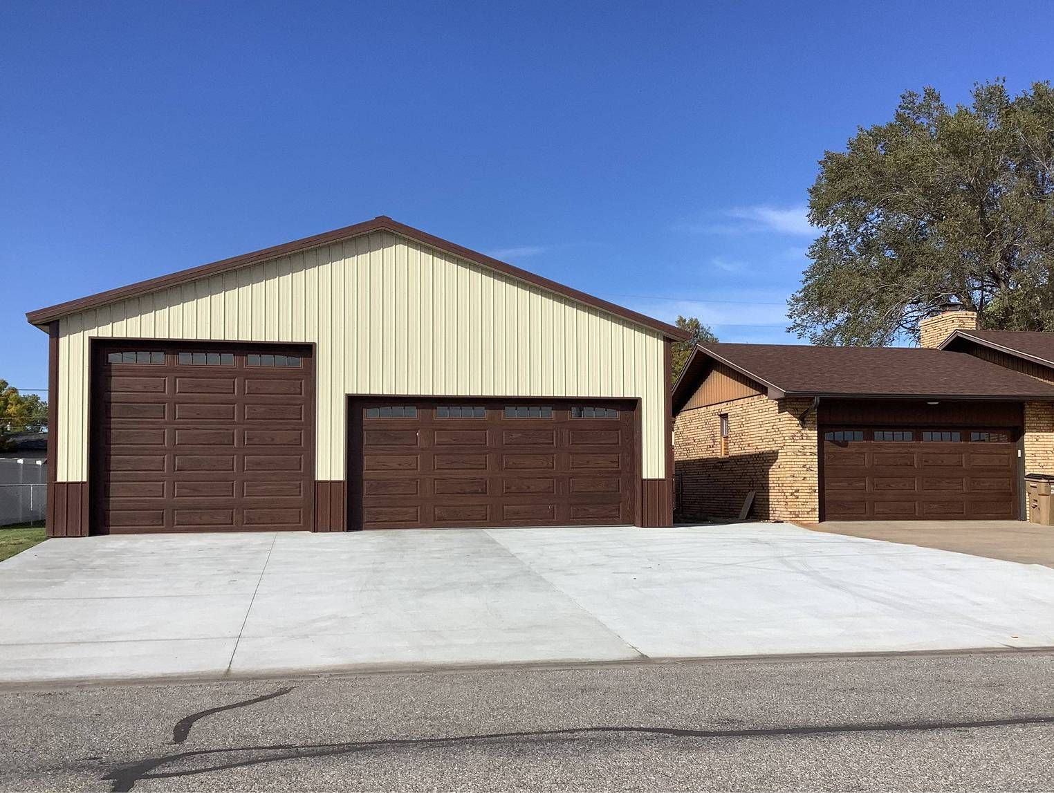 Tan metal garage with two brown garage doors, connected to a brown roofed structure and concrete driveway.
