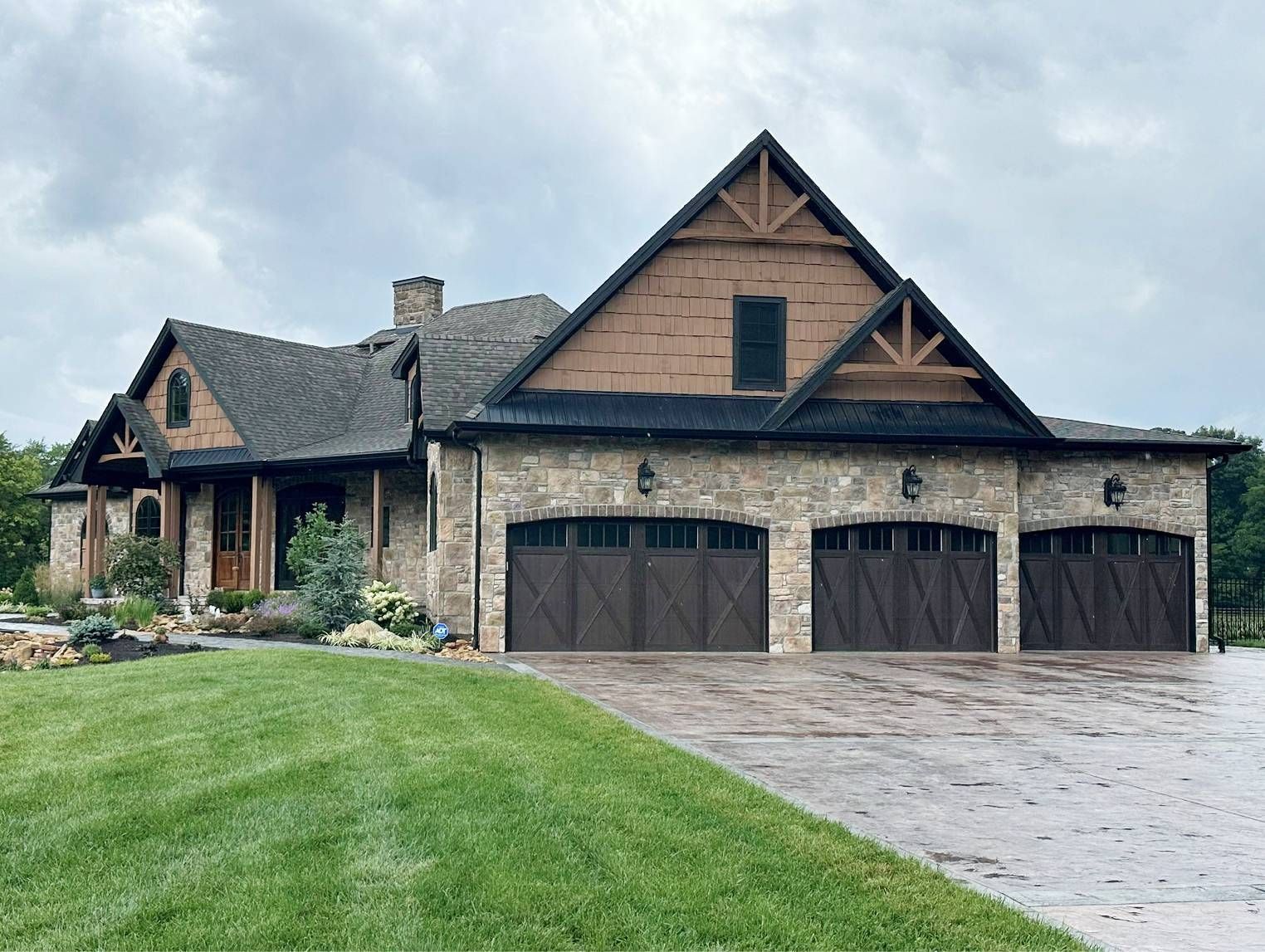 Stone and wood-accented home with three-car garage. Brown garage doors, green lawn, cloudy sky.