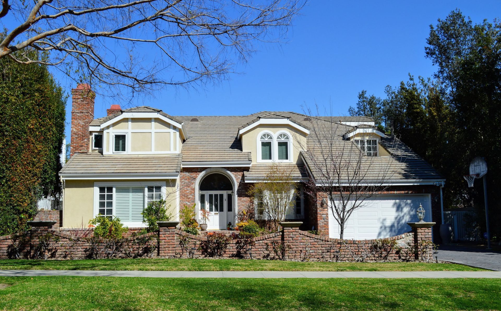 Two-story house with beige and red brick exterior, dormers, and a two-car garage under a blue sky.