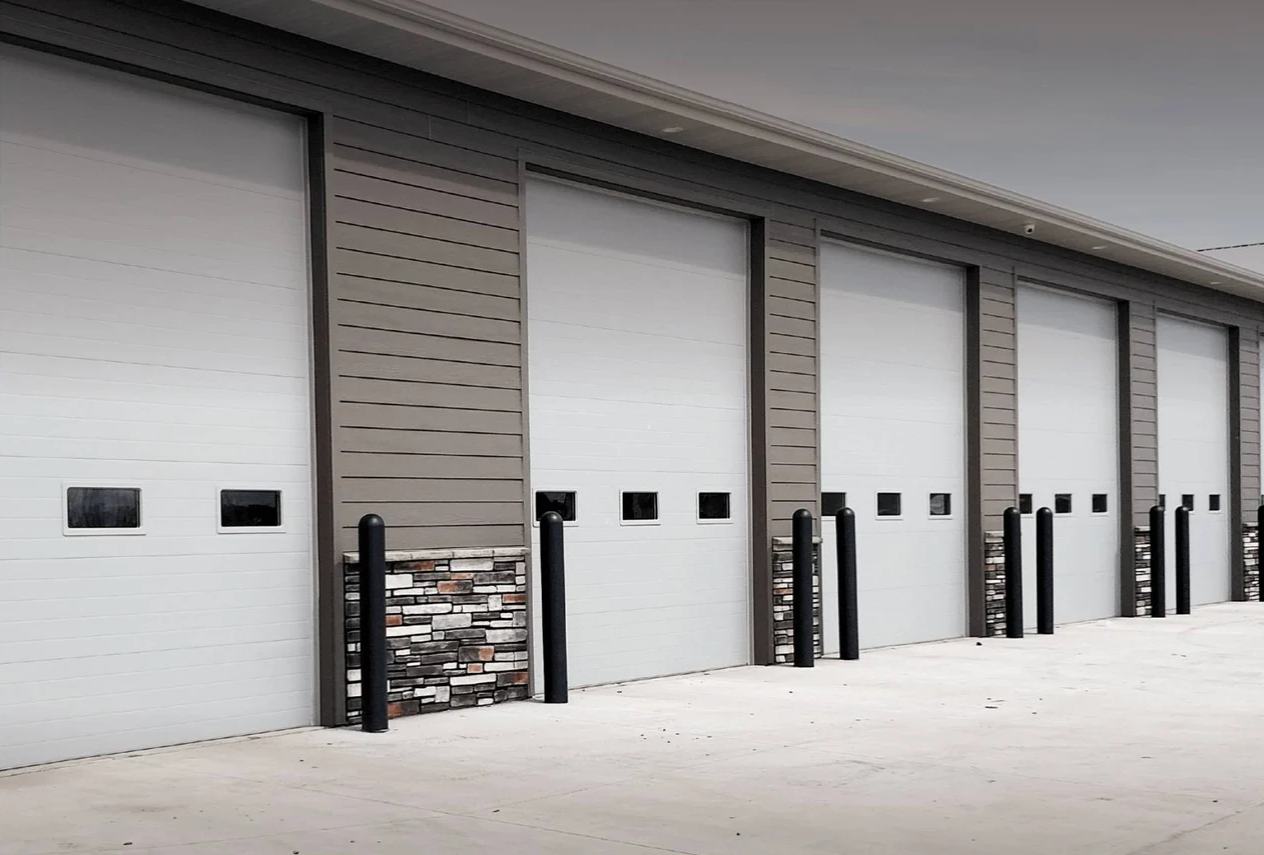 Exterior view of white garage doors in a row, with textured stone accents on the walls and black bollards in front.