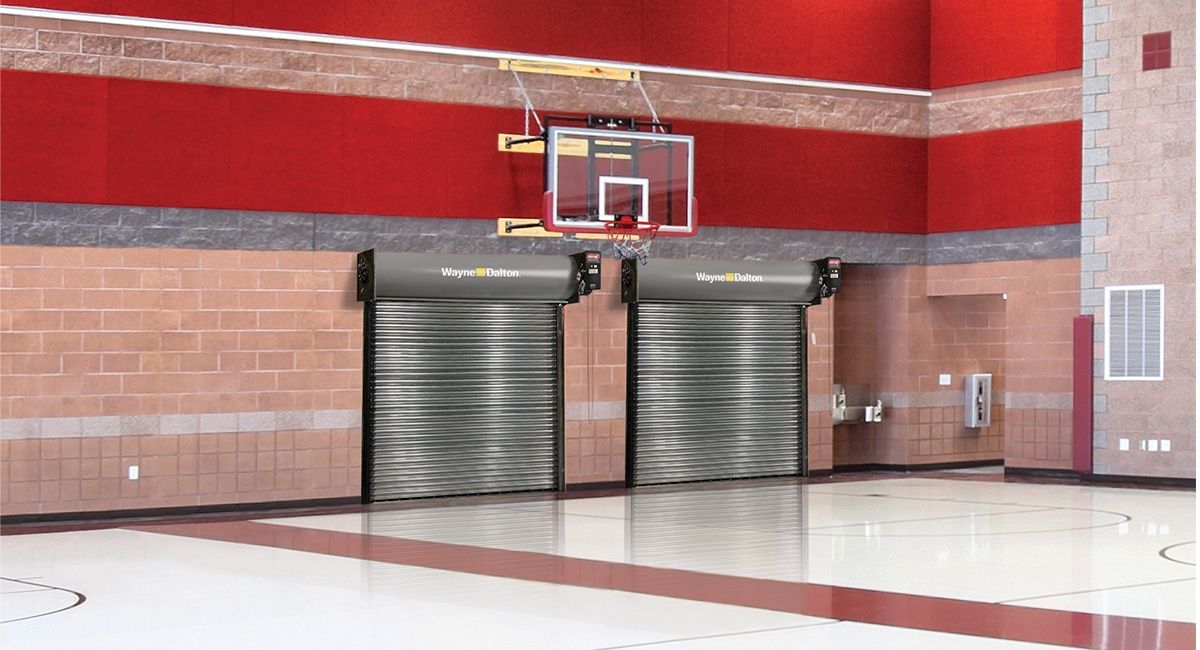 Basketball hoop mounted on a red wall above two closed, metal roll-up doors in a gymnasium.