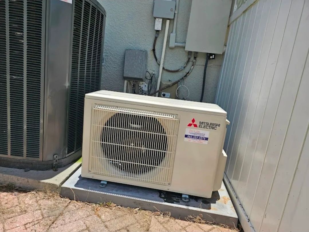 A Mitsubishi outdoor HVAC unit on a concrete slab next to a large air conditioner against a textured wall.