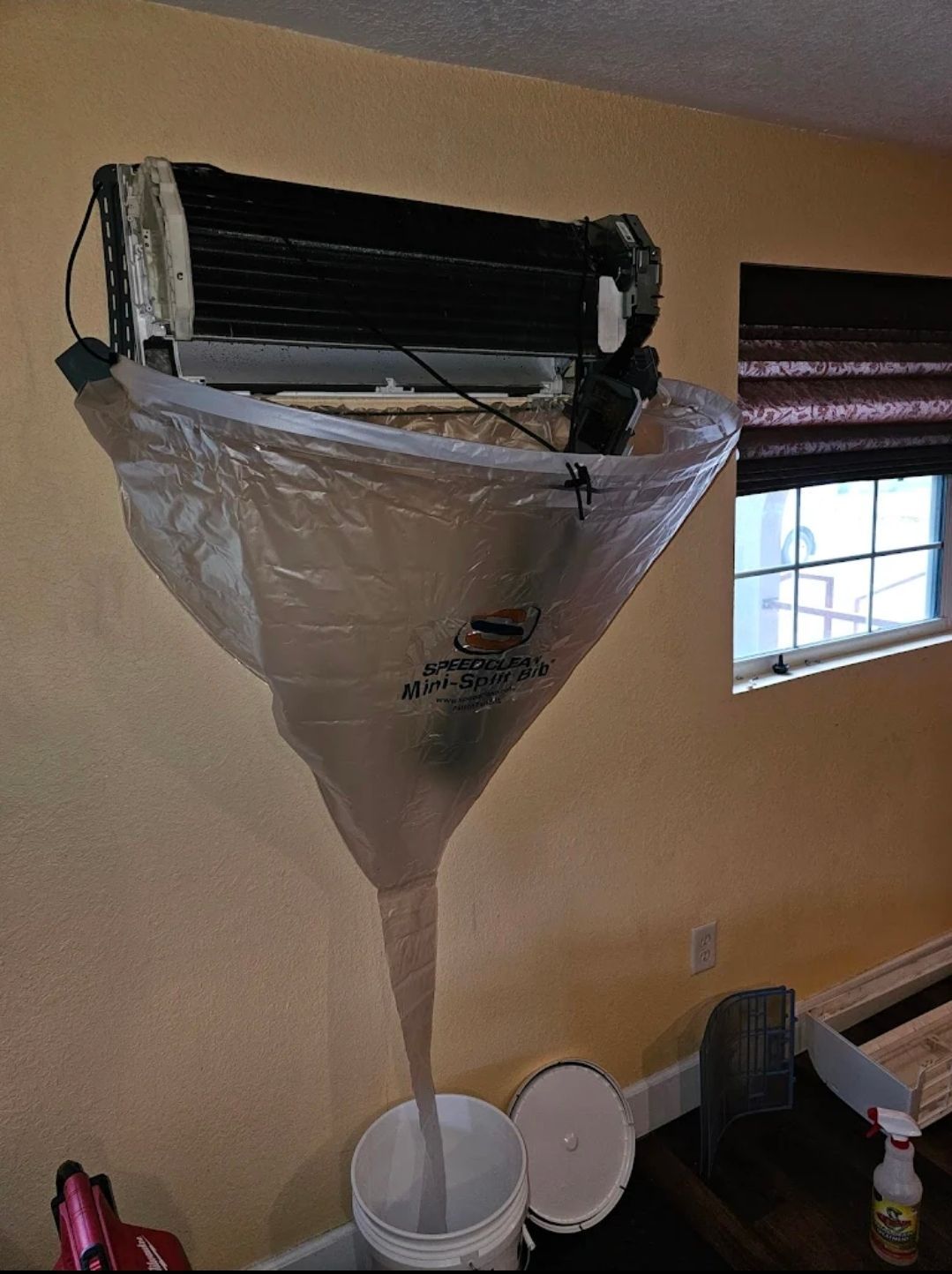 A mini-split air conditioner unit is being deep cleaned with a specialized funnel bag draining into a bucket below.