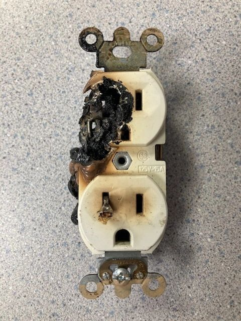 A burnt electrical outlet with heavy charring on the left side, mounted on a speckled surface.