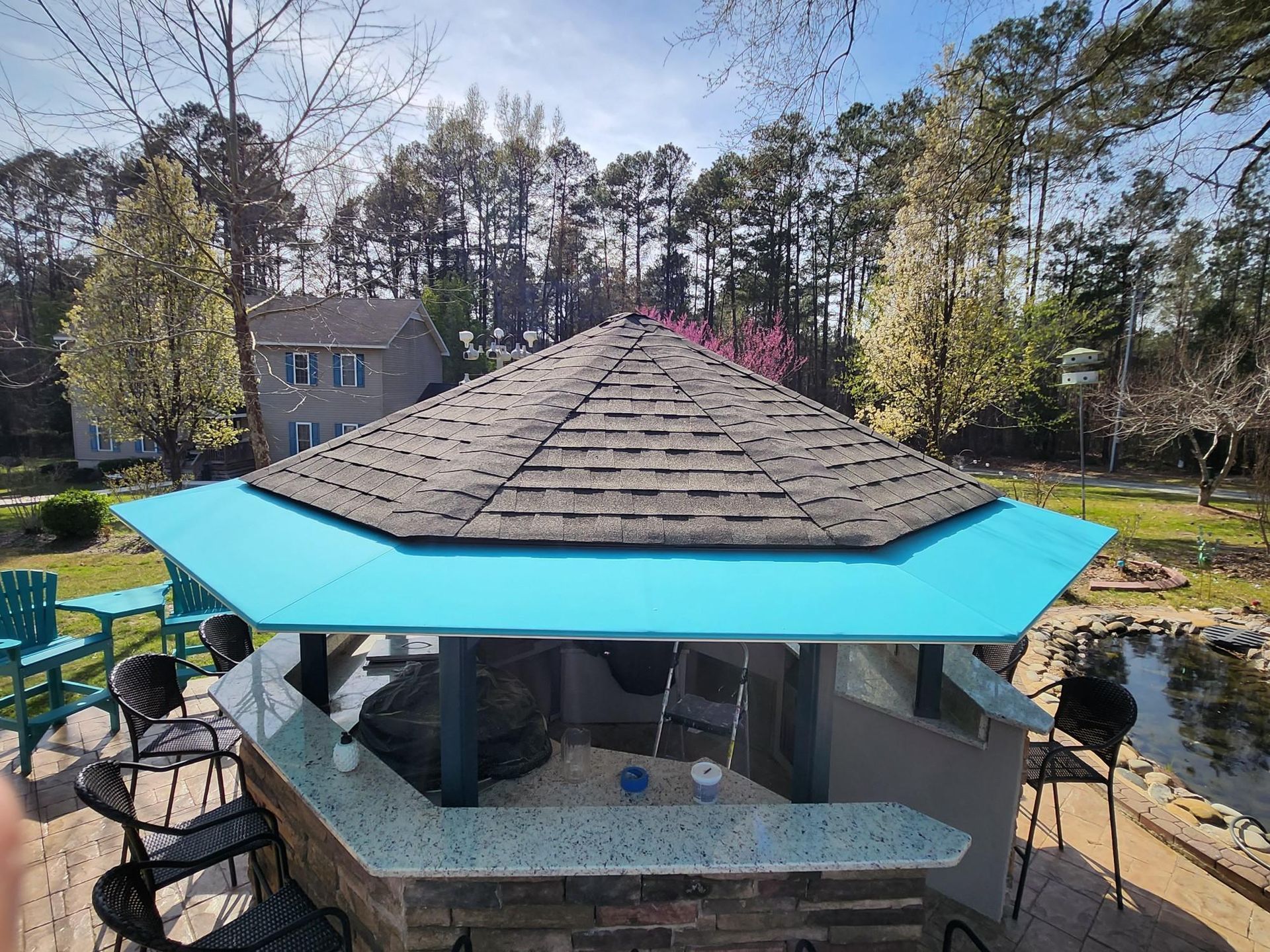 Backyard gazebo with blue roof and stone counter beside a pond, surrounded by trees and chairs