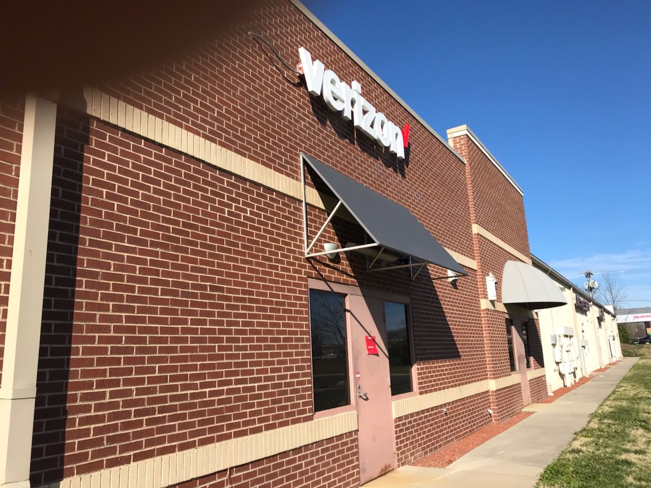 Brick storefront with a black awning and sign, viewed along a sidewalk on a sunny day