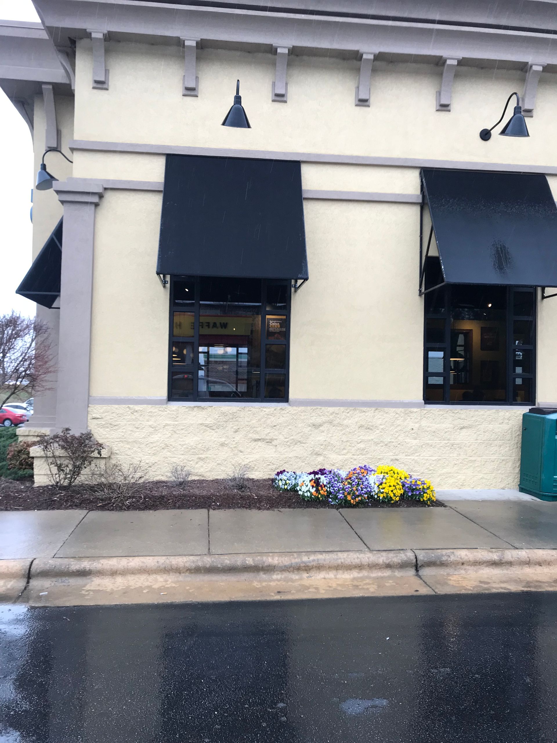 Storefront with black awnings, beige exterior, flower bed, and wet sidewalk in front.