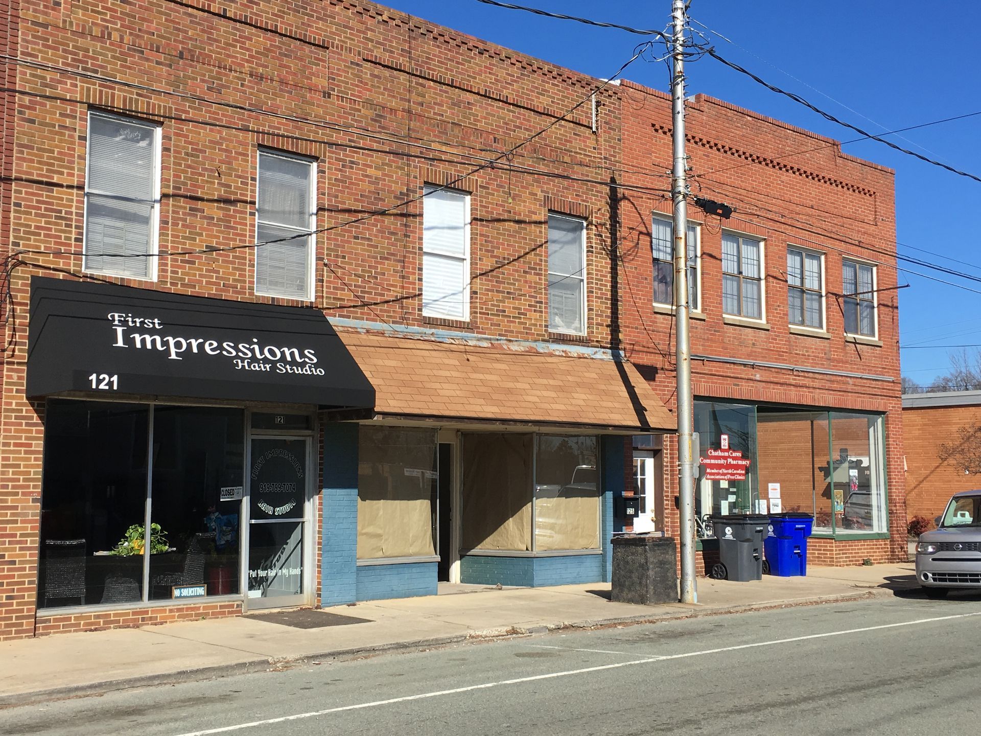 Brick storefronts on a sunny street, including an “Impression” sign and a blue recycling bin