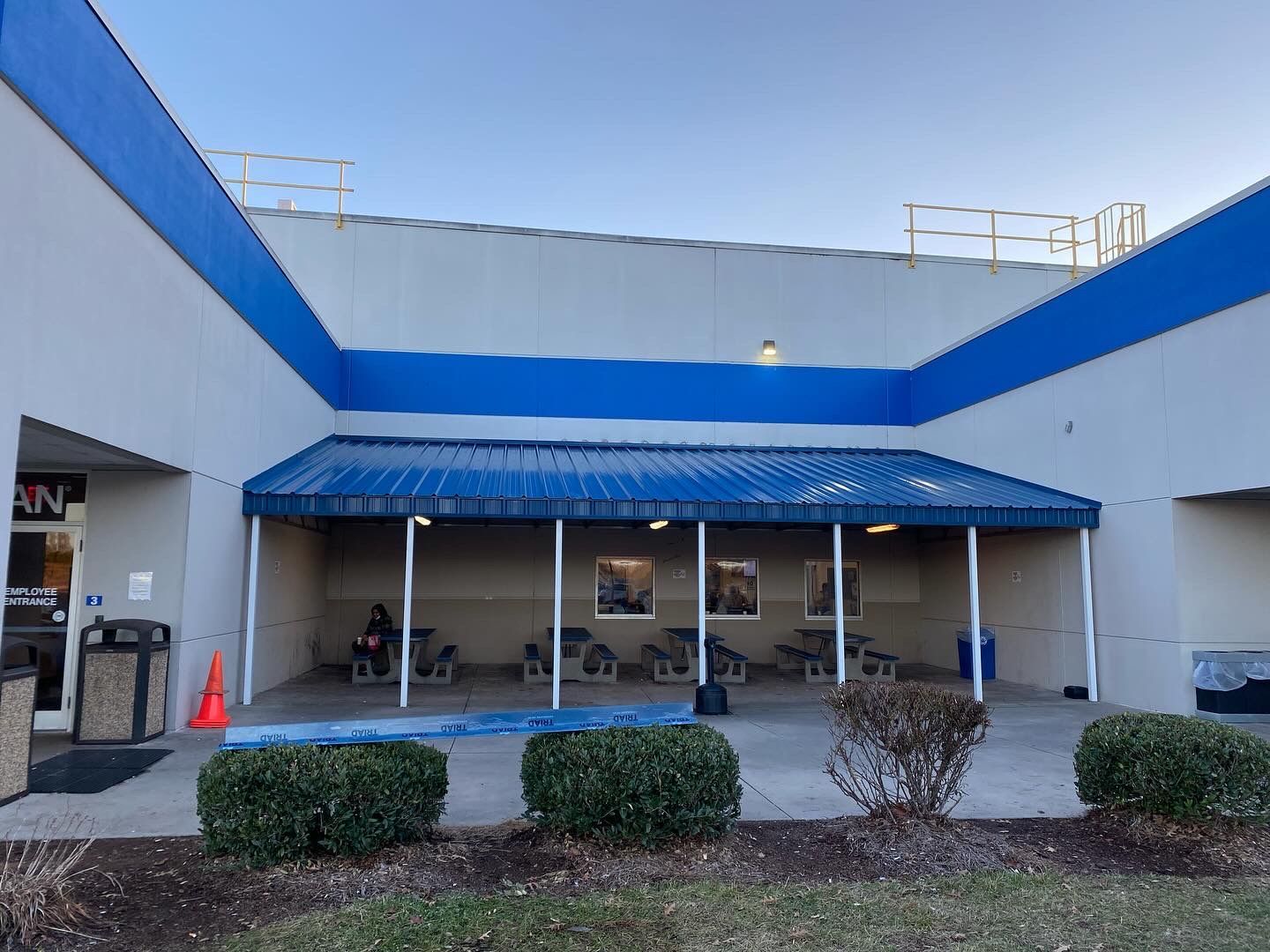 Storefront with blue awning and white walls, facing a small courtyard with shrubs and an open entrance.