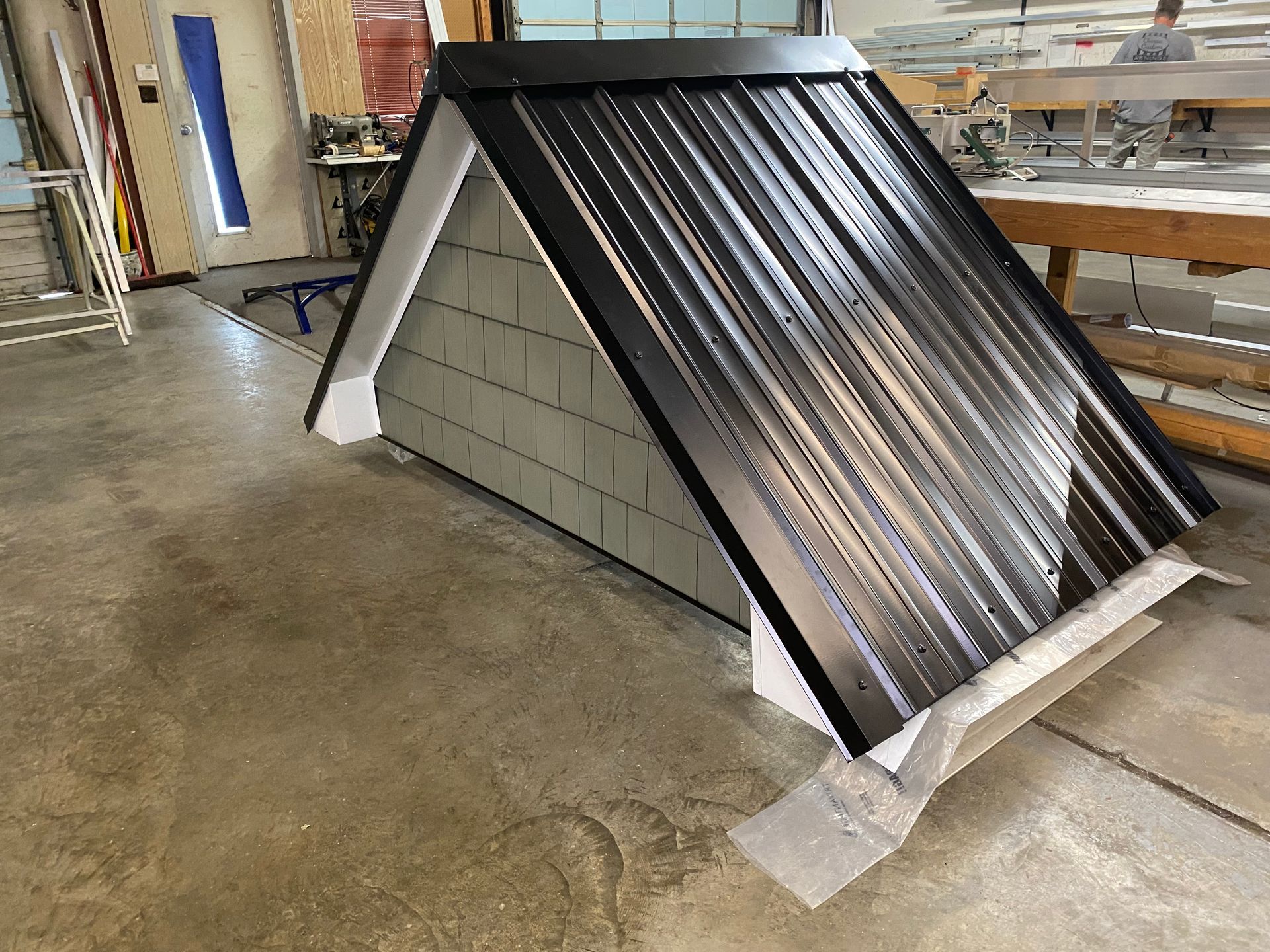 Small A-frame roof model with black metal panels in a workshop, on a concrete floor.