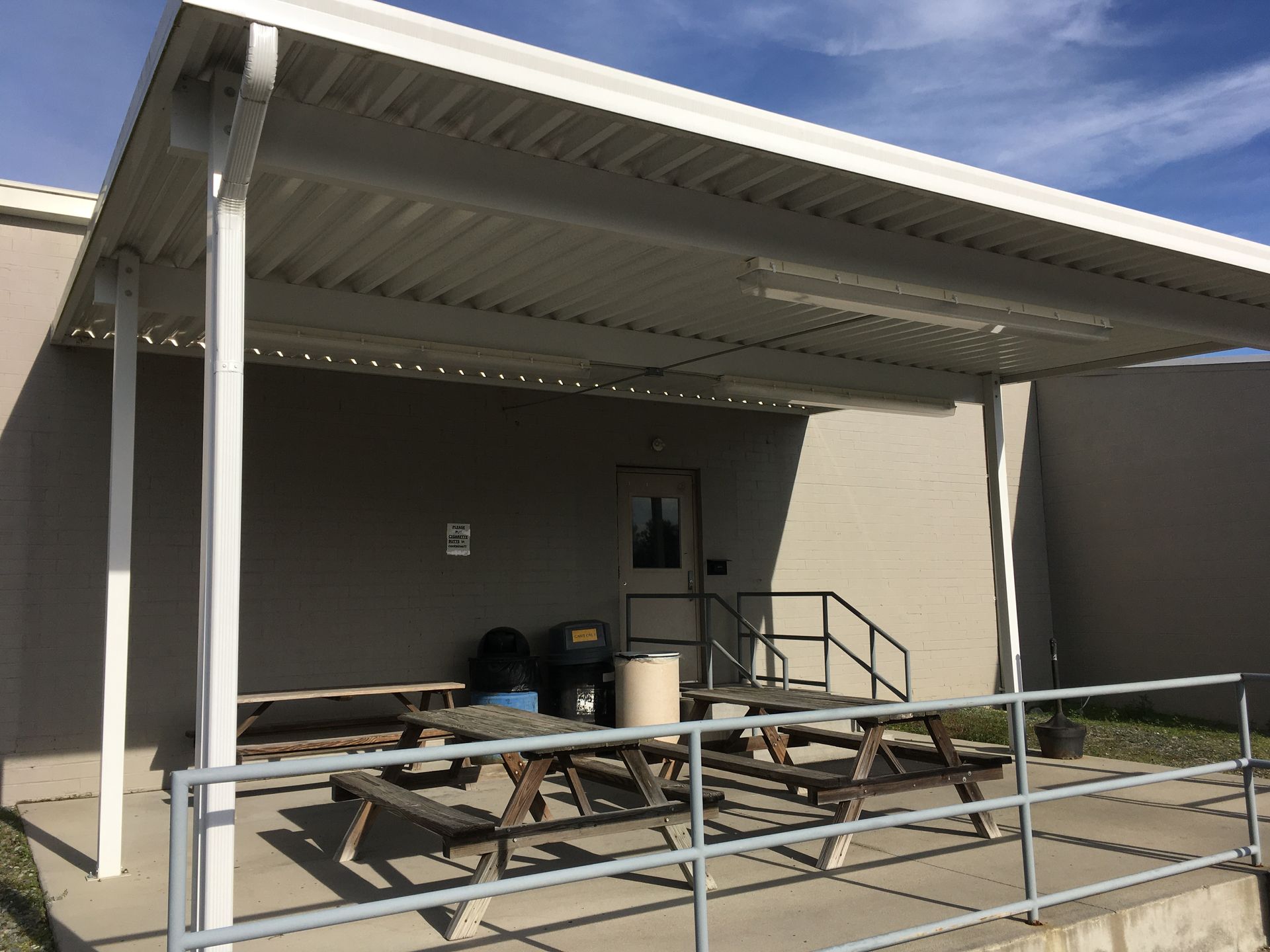 Covered outdoor patio with picnic tables, metal railing, and a ramp beside a building under a blue sky
