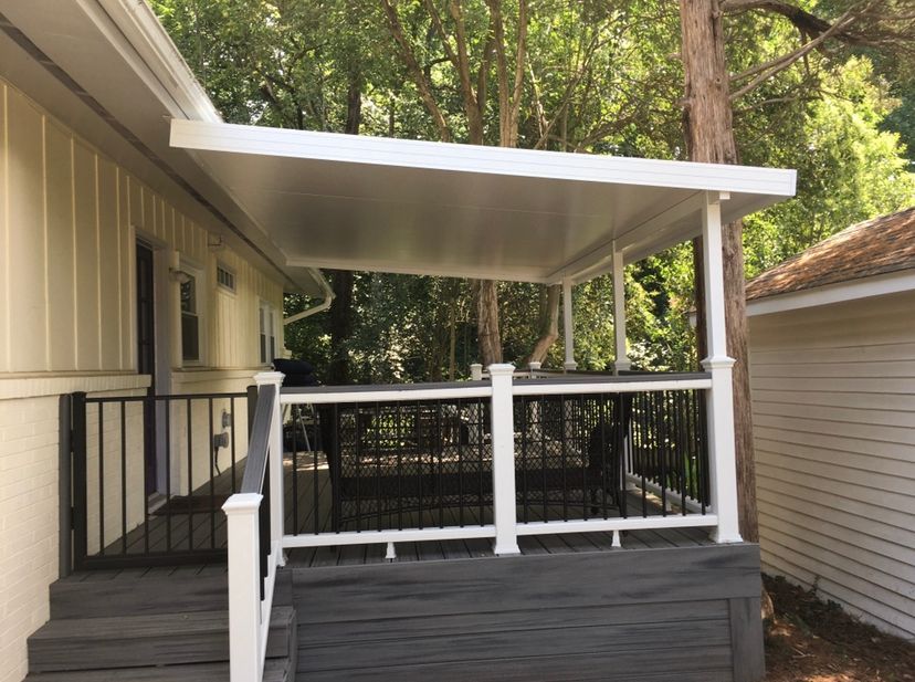 Covered porch with white railing and dark skirting beside a house, backed by trees and a shed.