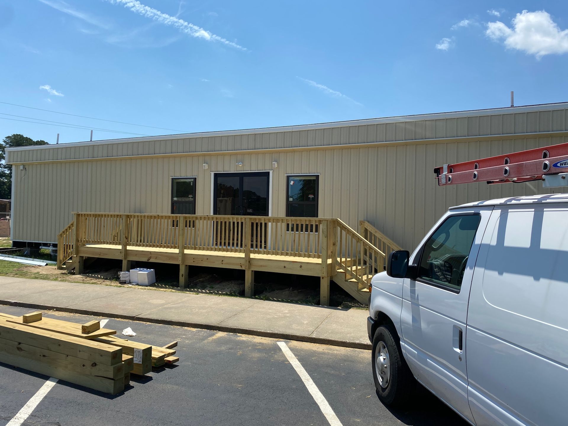 Portable office trailer with a wooden ramp and stairs, parked beside a white van in a sunny lot