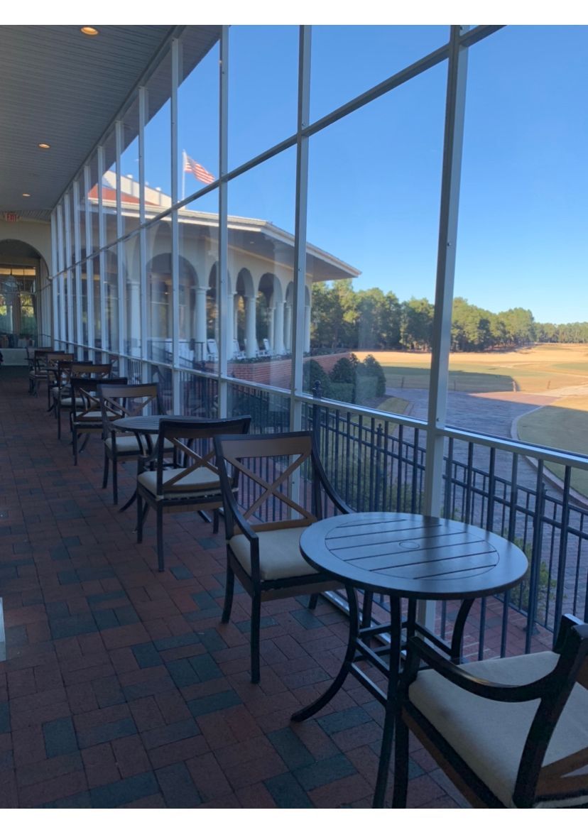 Covered patio with black metal tables and chairs overlooking a sunny riverside building and lawn