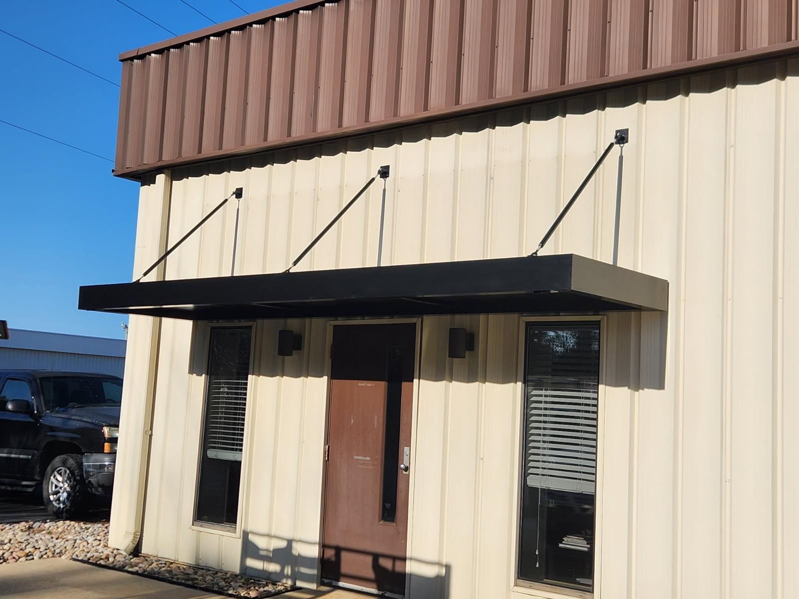 Small beige building facade with a dark awning, brown door, and two narrow windows under a blue sky