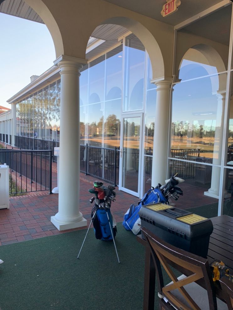 Golf bags on a covered patio beside arched columns and a large windowed building.