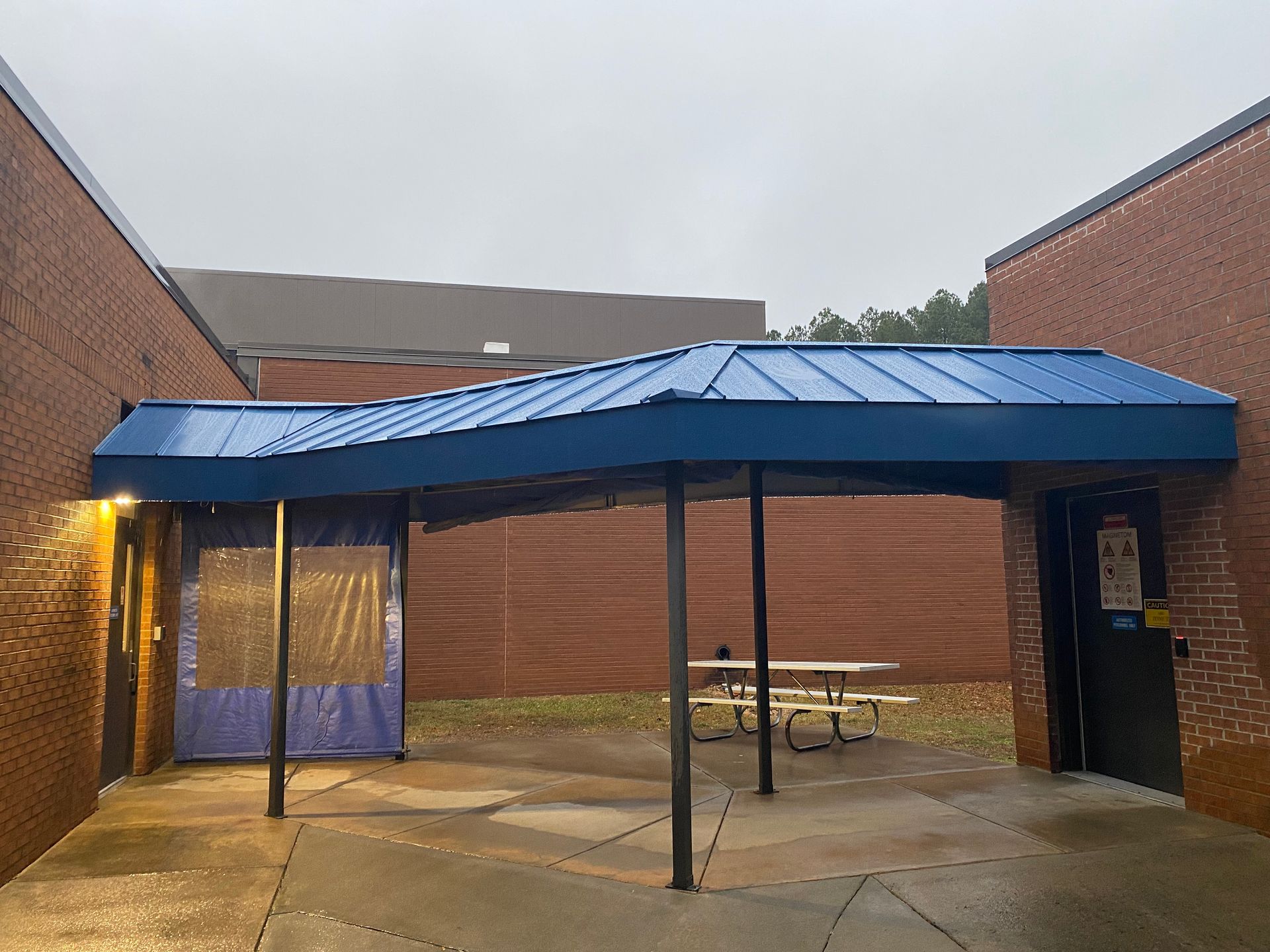 Covered courtyard with blue metal canopy between brick buildings, wet concrete ground, and picnic tables.