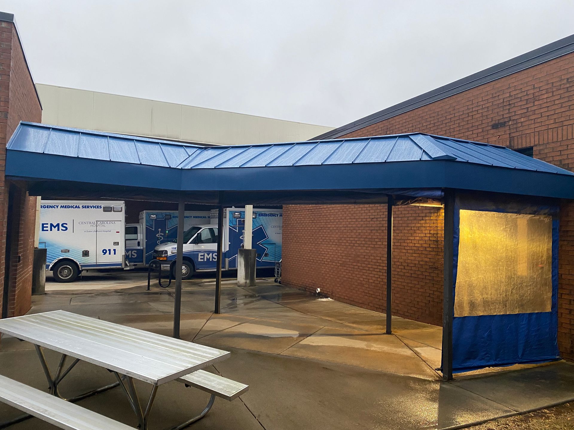 Covered outdoor walkway beside a brick building, with picnic tables and a parked van in the background.