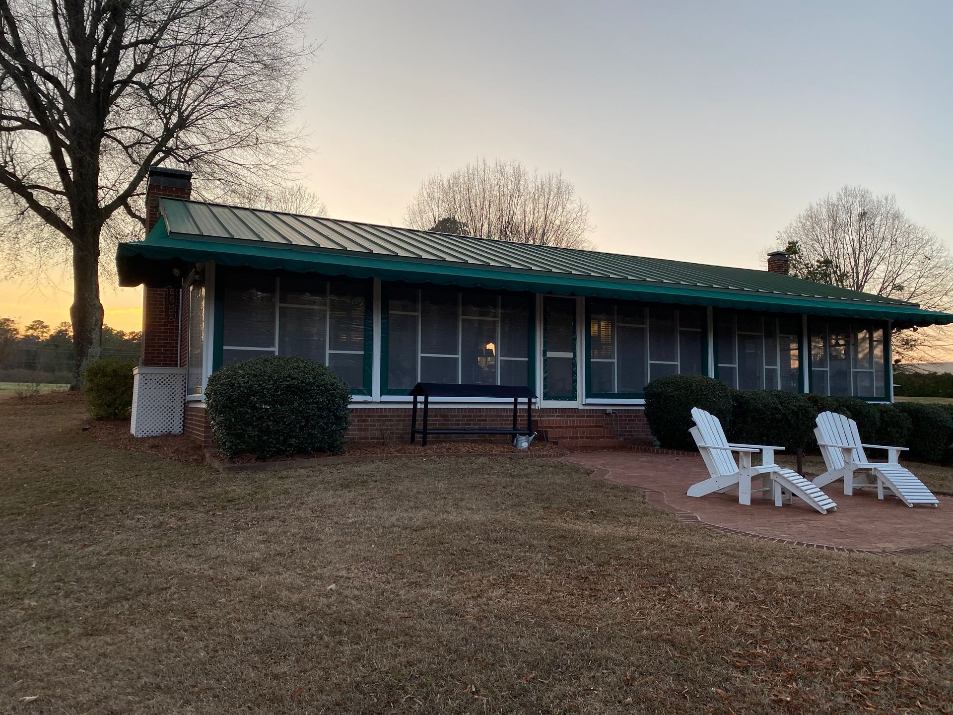 Small cabin with green roof at dusk, framed by trees and lawn chairs on the front porch.