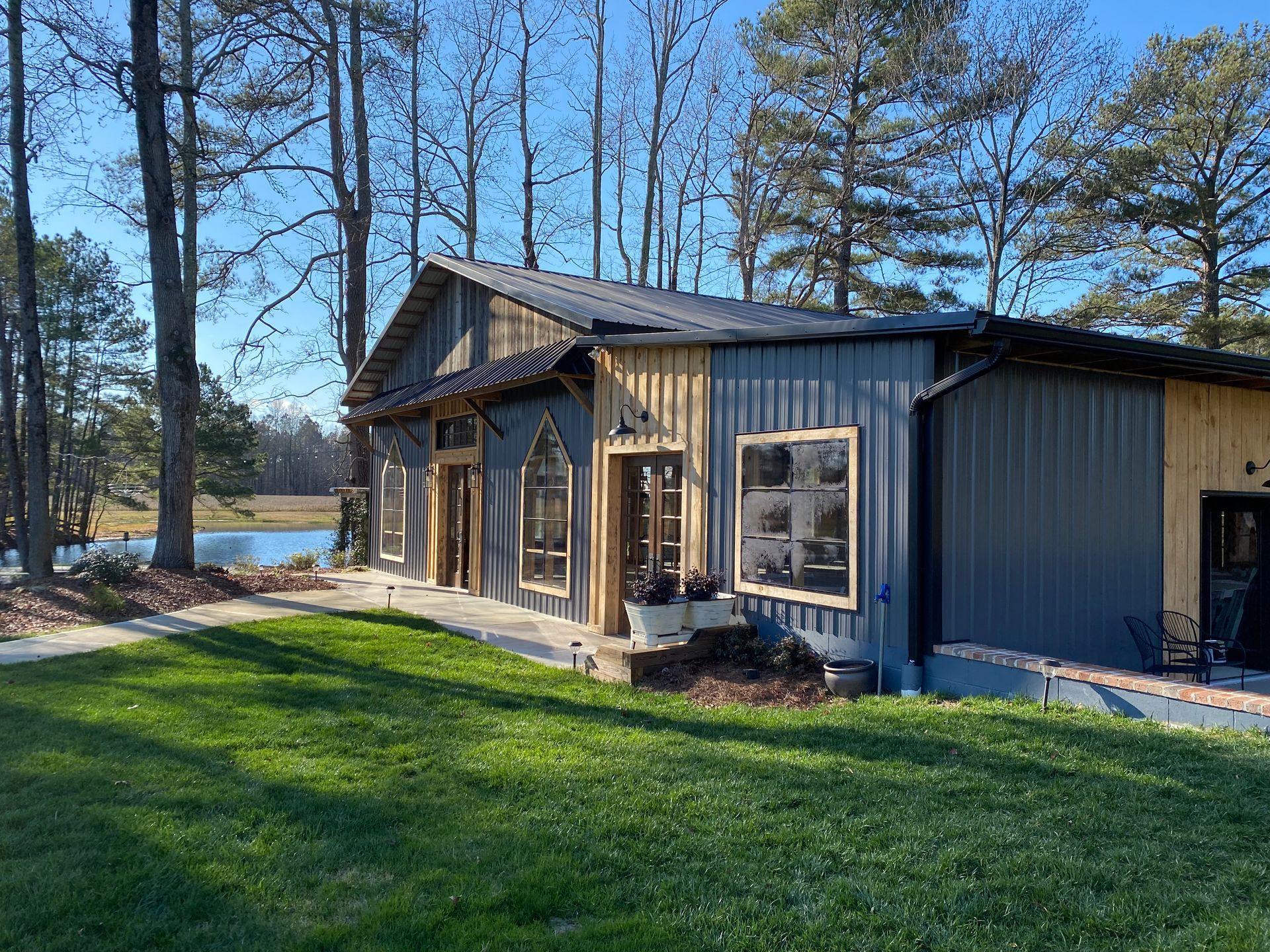 Small gray cabin with a porch beside a green lawn and lake, surrounded by tall pine trees.