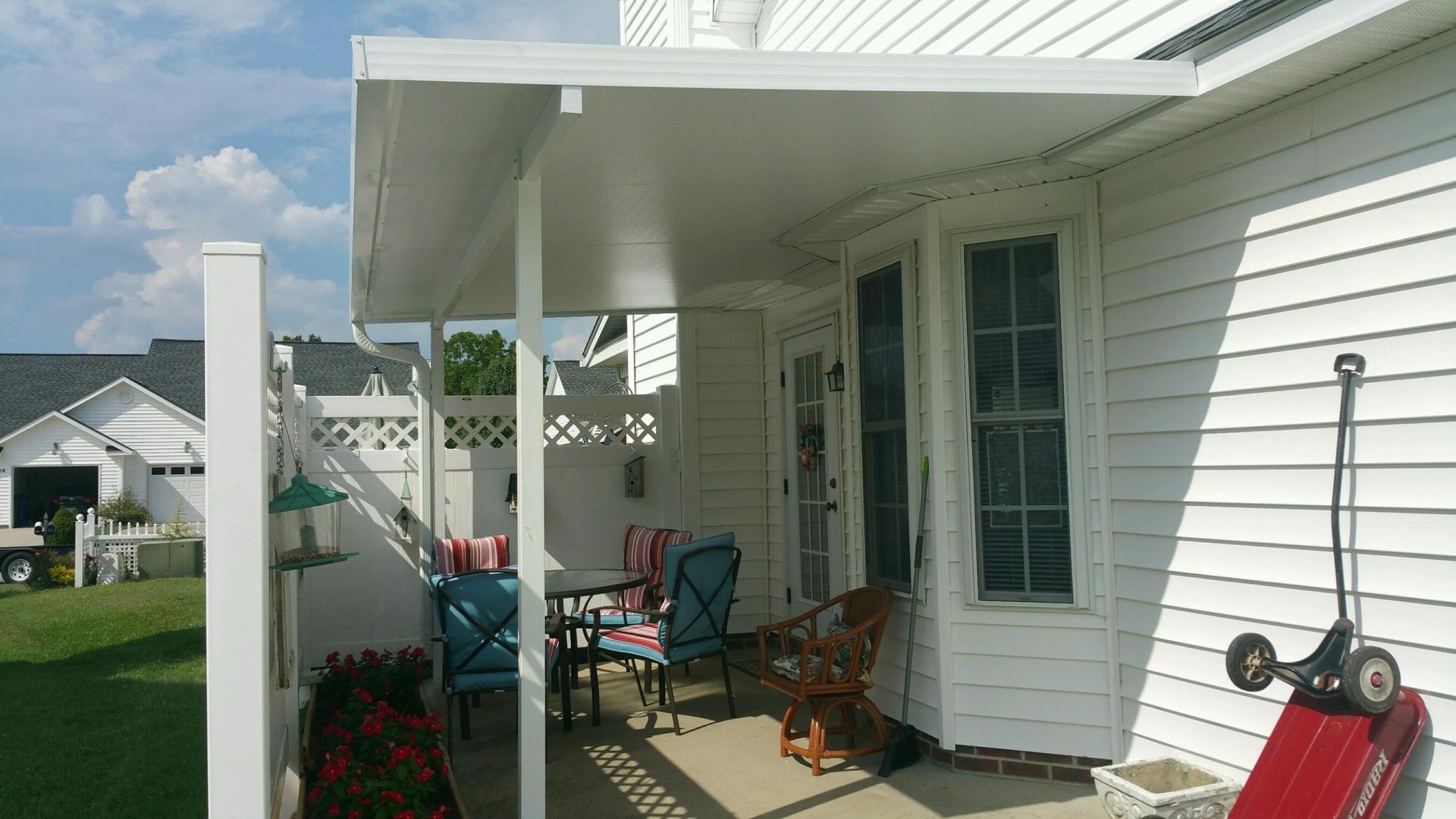 White house patio with covered porch, teal chairs, grill, potted flowers, and a red wagon on the right
