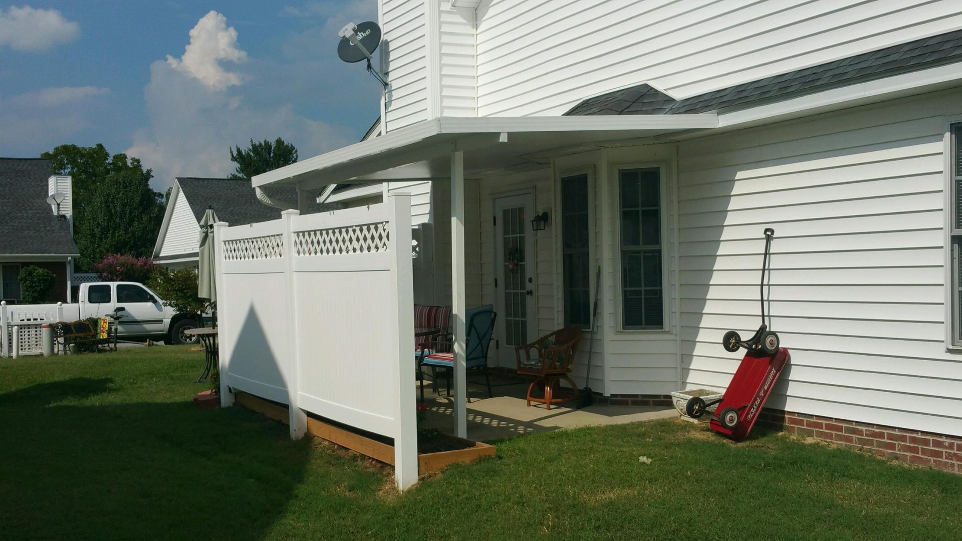 White vinyl house patio with a white fence, porch chairs, and a red lawn mower on green grass.