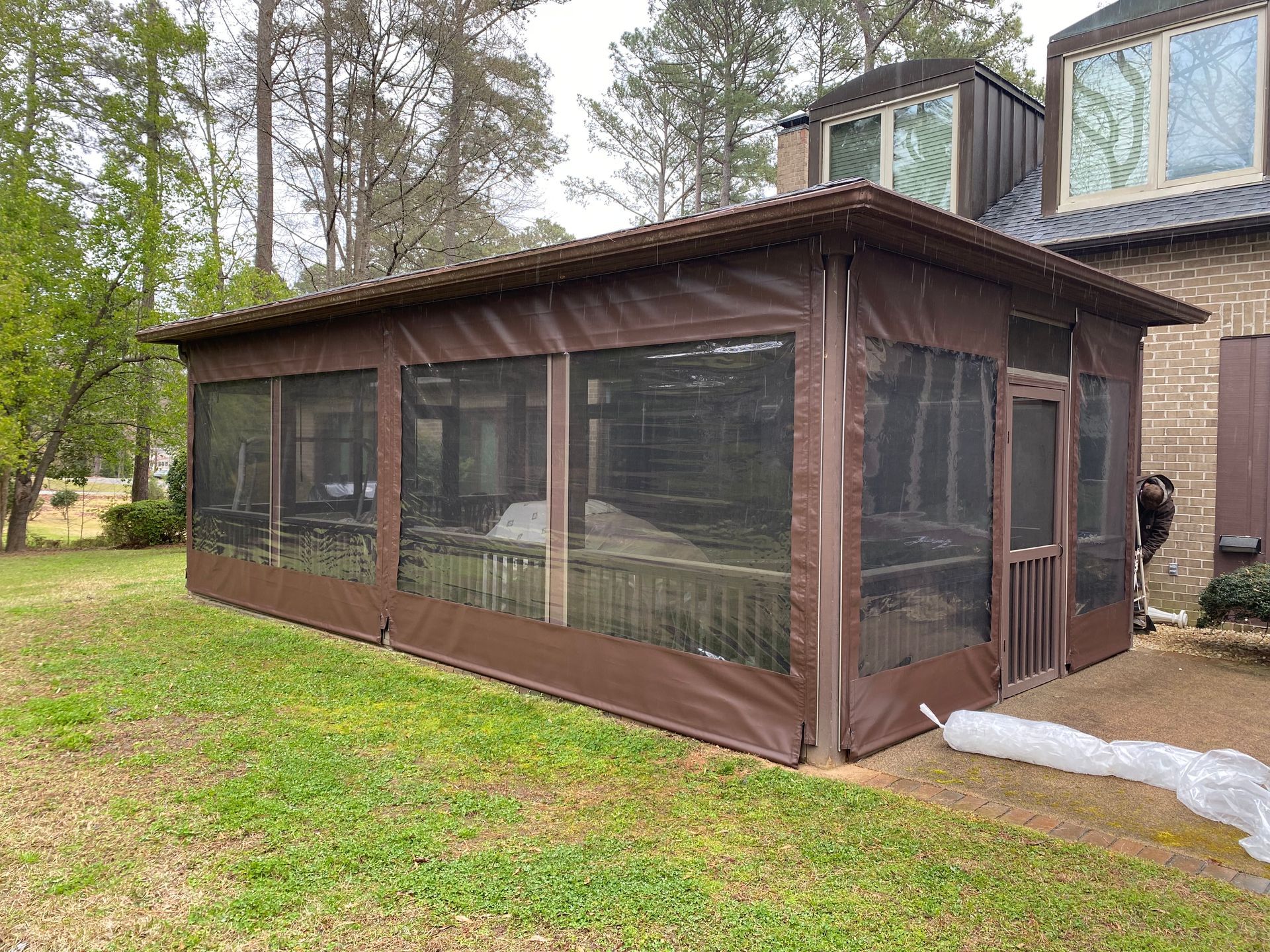 Brown screened porch attached to a house in a grassy yard with trees around it