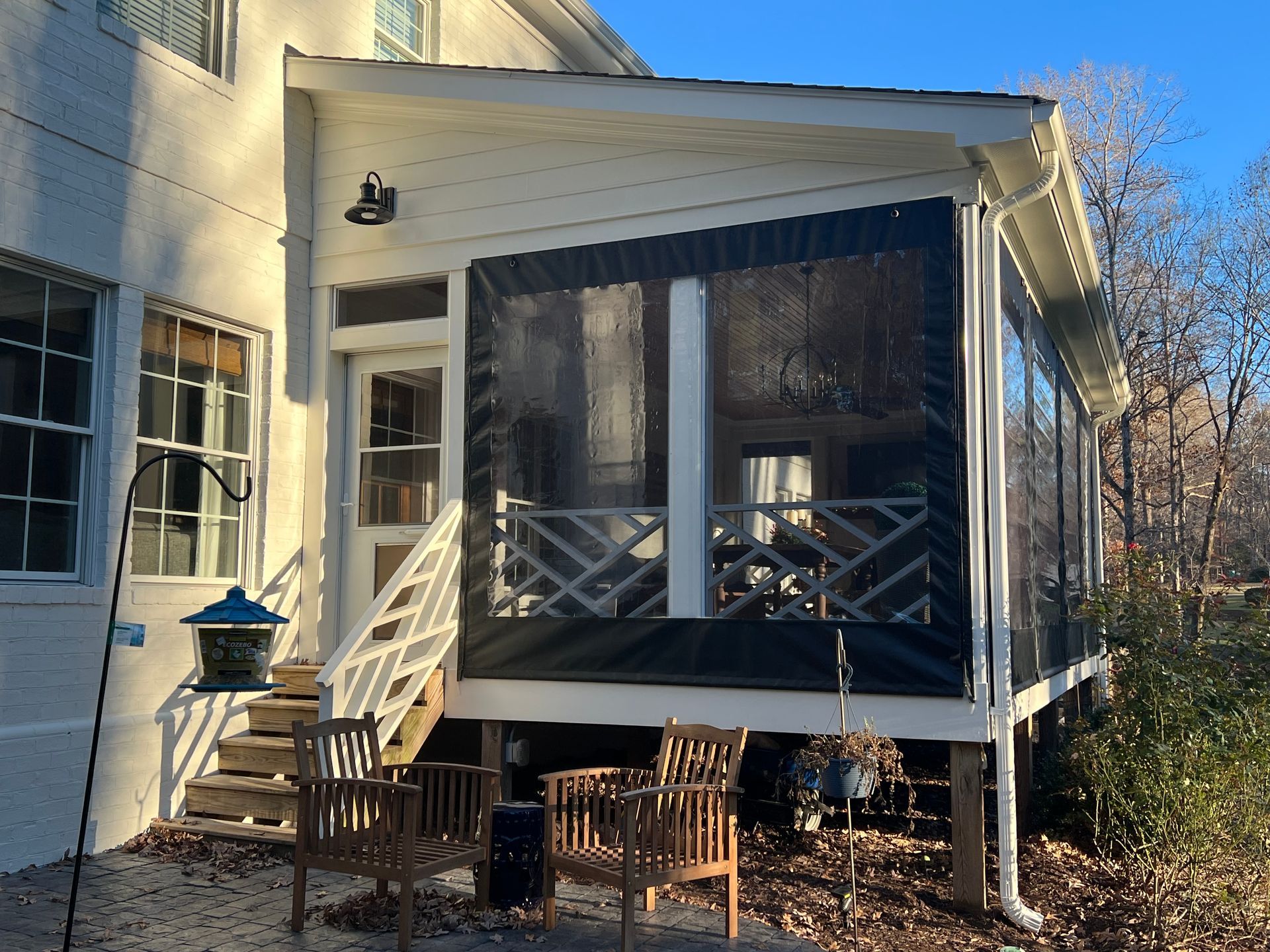 Covered porch with screened windows, white railings, and wooden chairs beside a house in sunlight
