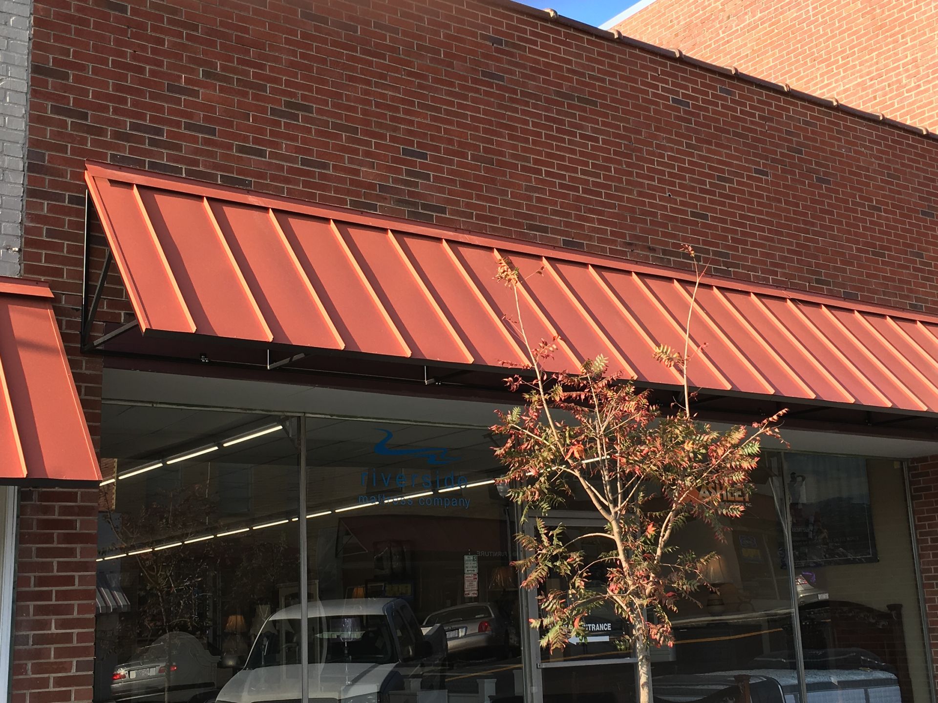Brick storefront with red metal awning and large glass windows, with a small tree in front.