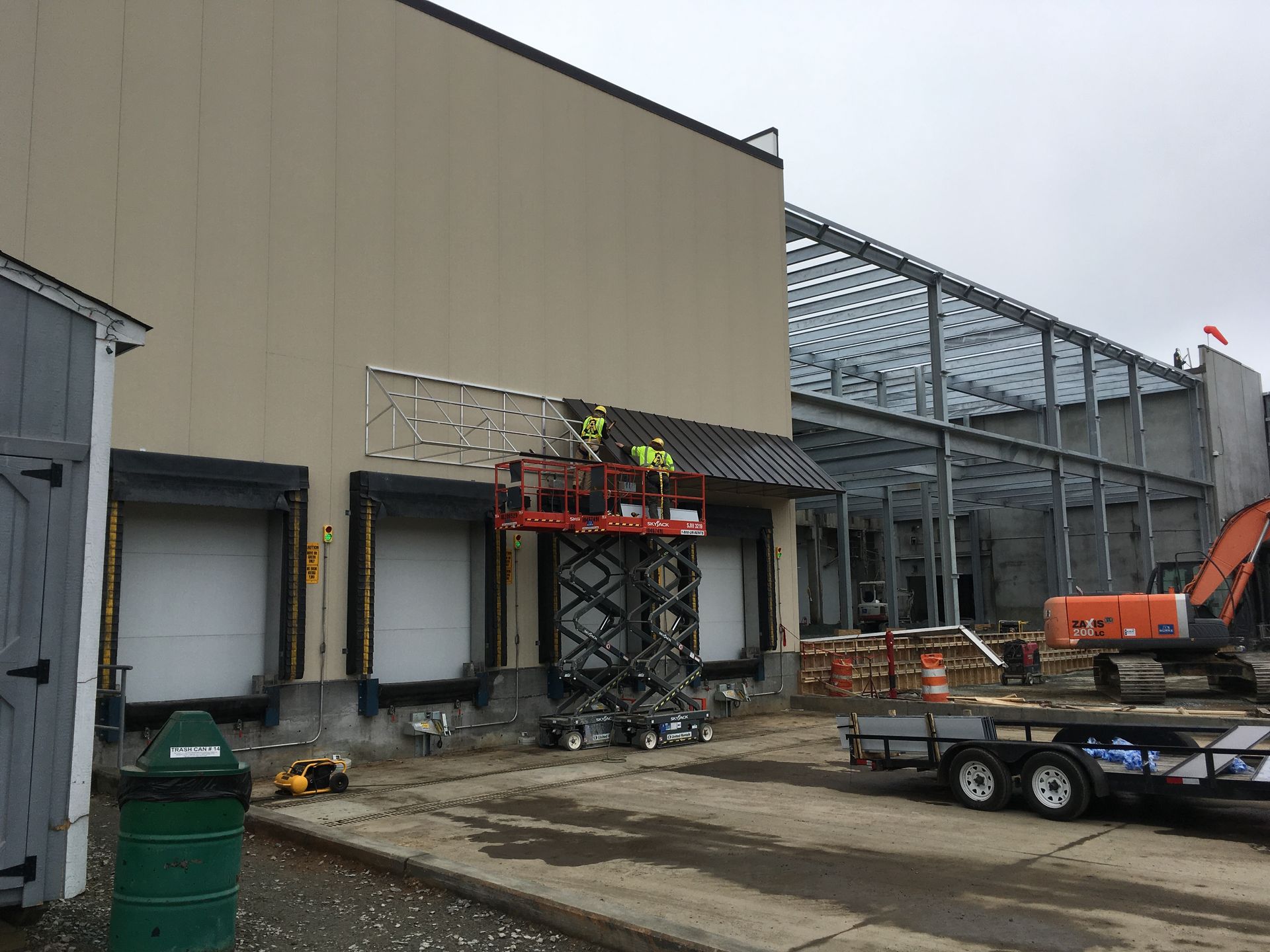 Construction site outside a beige industrial building with scaffolding, lifts, and a trailer in front
