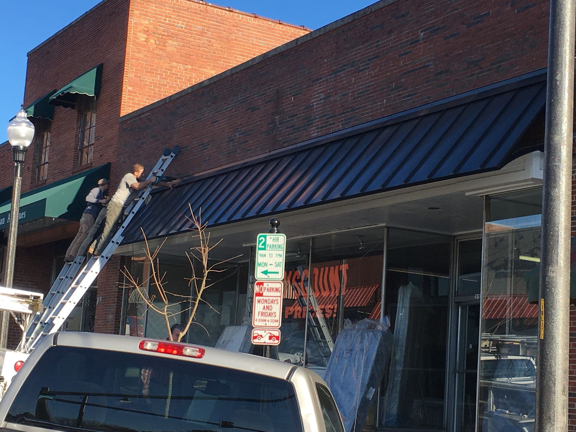 Workers on a ladder repairing a storefront awning above parked cars on a city street