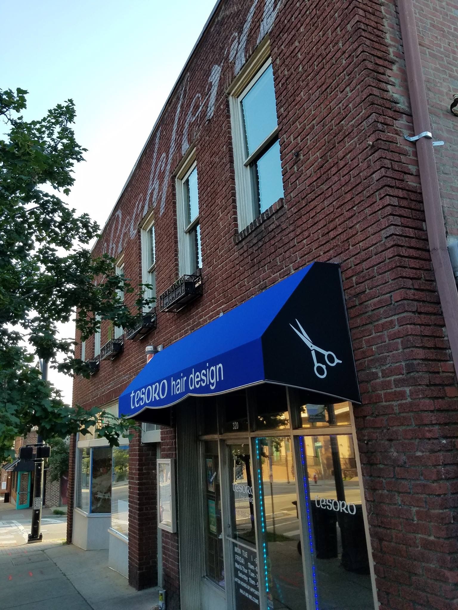 Brick storefront with blue awning on a sunny street corner, next to a tree and sidewalk.