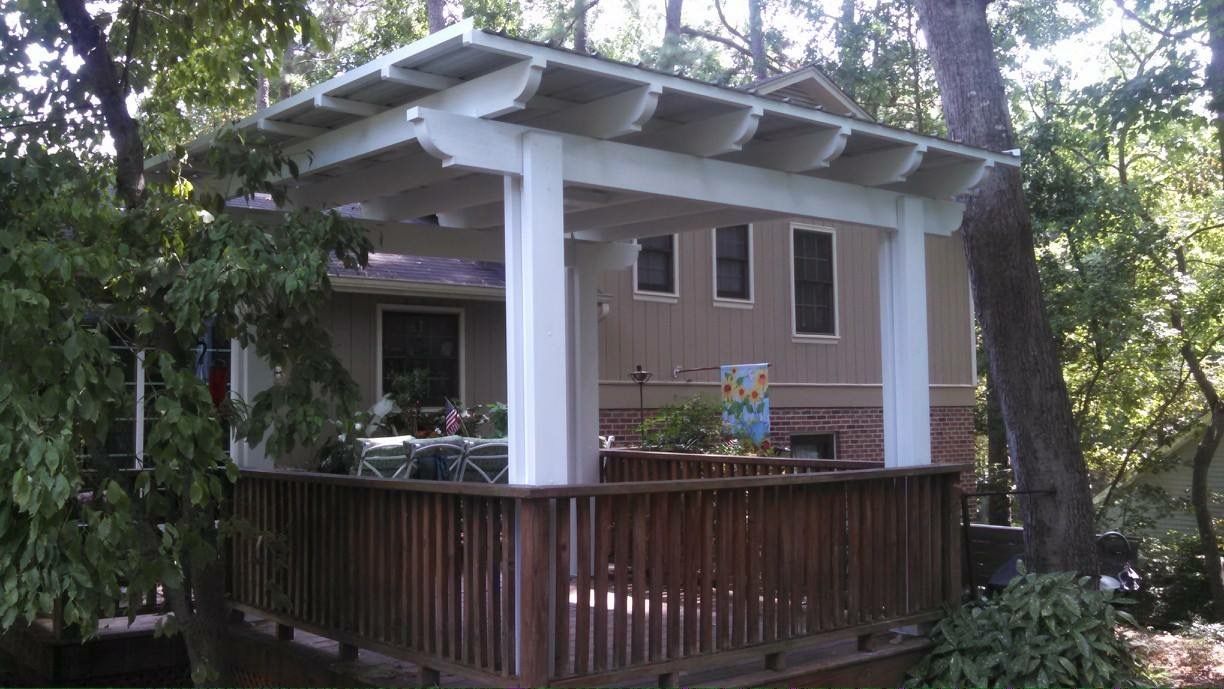 Wooden deck with pergola beside a beige house, surrounded by trees and greenery